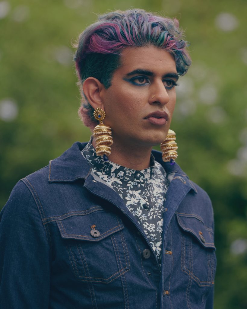 Alok Vaid-Menon in a denim jacket, from a 3/4 view. Their dark hair is streaked with red and brushed high away from their face. They're wearing heavy gold earrings and a floral shirt.