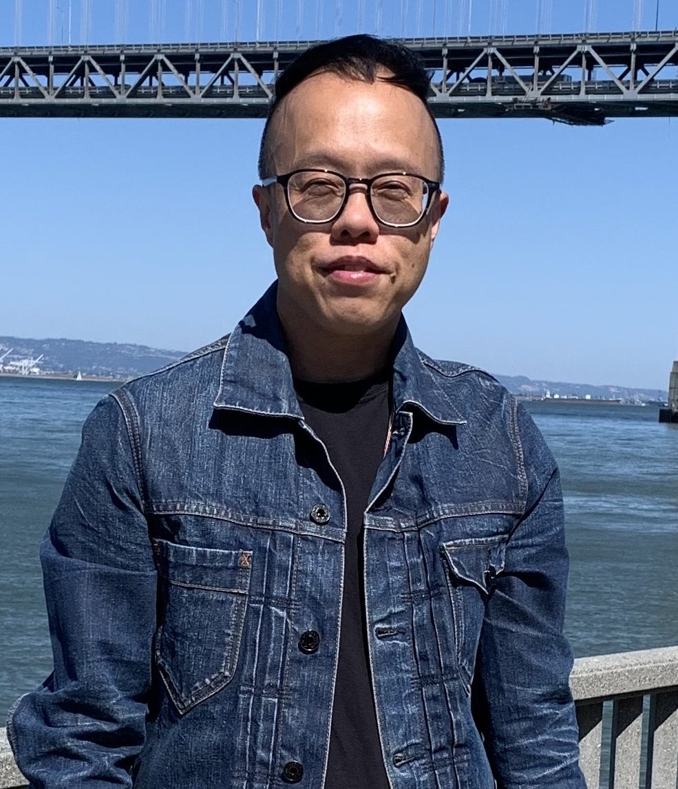 Jian Neo Chen in a dark denim jacket, standing in front of a bridge and a large body of water. 