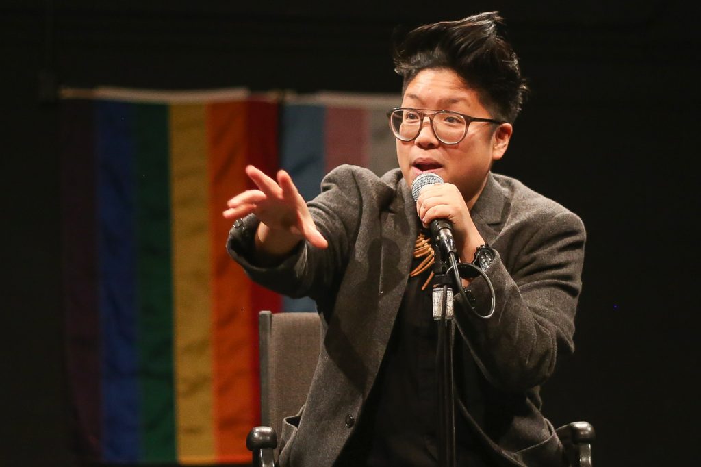 Kay Ulanday Barrett in a tan blazer, one hand on a microphone, another reaching out toward the audience. They're on a stage, speaking animatedly. 