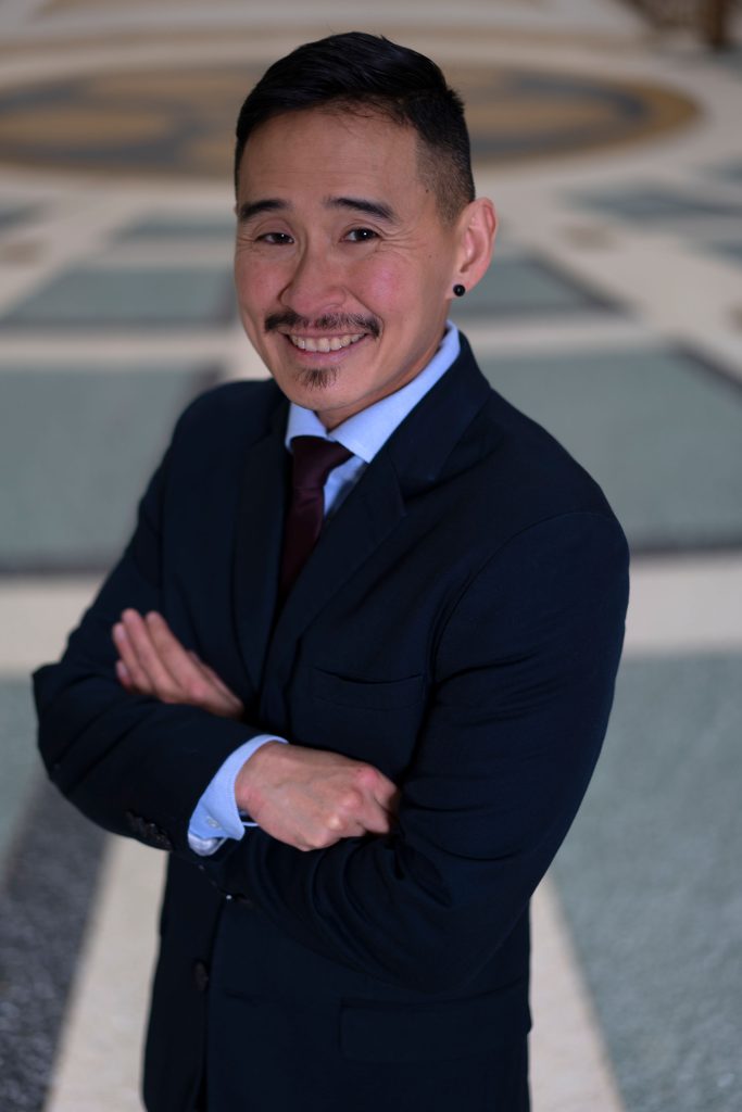 Kris Hayashi in a dark suit with a blue shirt and red tie. He stands with his arms crossed, smiling at the camera.