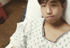 Jo, a trans nonbinary Taiwanese American, in a hospital bed and gown with an IV in their right arm