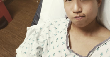 Jo, a trans nonbinary Taiwanese American, in a hospital bed and gown with an IV in their right arm