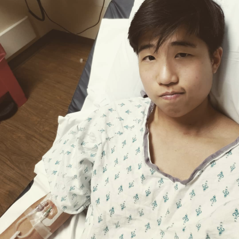 Jo, a trans nonbinary Taiwanese American, in a hospital bed and gown with an IV in their right arm