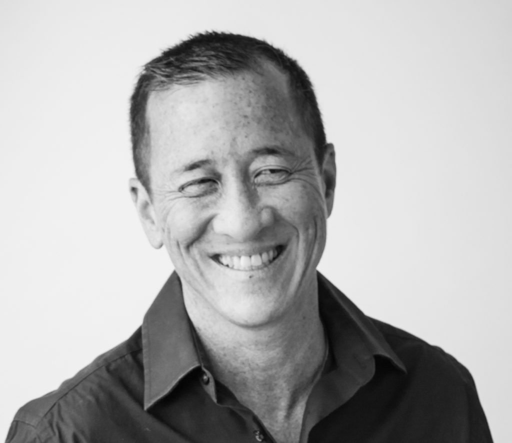 A black and white photo of Willy Wilkinson. He wears a dark button-up shirt and is smiling at the camera, before a white backdrop. 