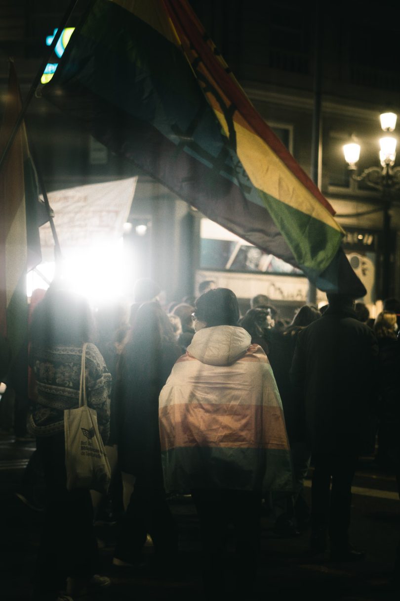 A photo of a crowd at night. A person wrapped in a transgender pride flag is at the center. That flag and a queer pride flag hanging over the top are the only spots of color in an otherwise grayscale photograph