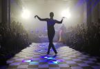 a ballroom dancer photographed from behind on a checkered floor. Their arms are raised to either side and their legs crossed in a still pose.