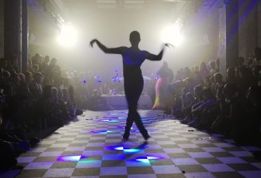a ballroom dancer photographed from behind on a checkered floor. Their arms are raised to either side and their legs crossed in a still pose.