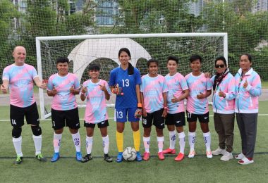 ten people in soccer uniforms standing on a field, in front of a goal. Nine of them are wearing pink and blue jerseys (in the colors of the trans flag), and one, in the center, is in a blue jersey and shorts with goalie gloves.